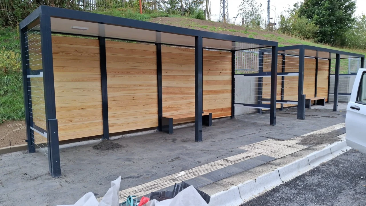 Zwei Wartehallen mit grünem Dach und Holzrückwand vor einem begrünten Hügel, neben einer Straße.