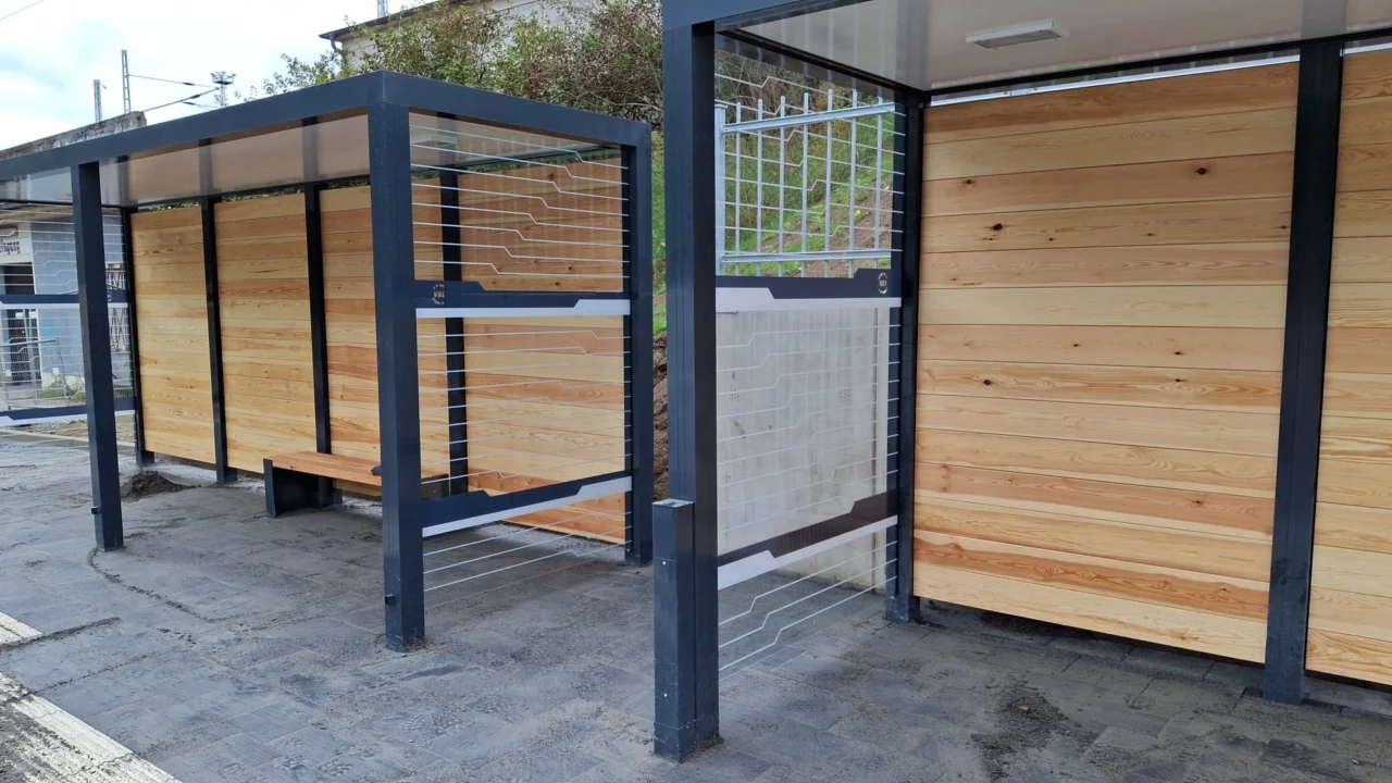 Zwei Wartehallen mit grünem Dach und Holzrückwand vor einem begrünten Hügel, neben einer Straße.