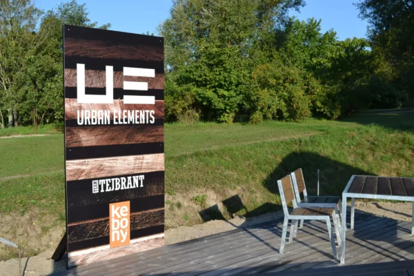 Schild mit Urban Elements und Team Tejbrant Logos vor gruener Wiese neben Stuehlen und einem Tisch.