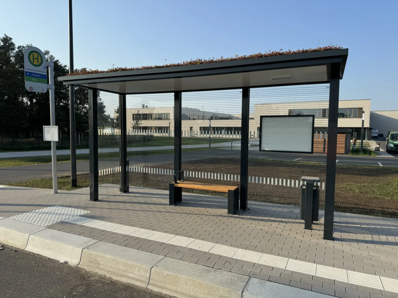 Wartehalle mit Gründach und Sitzbank an einer Bushaltestation vor einem größeren Gebäude.