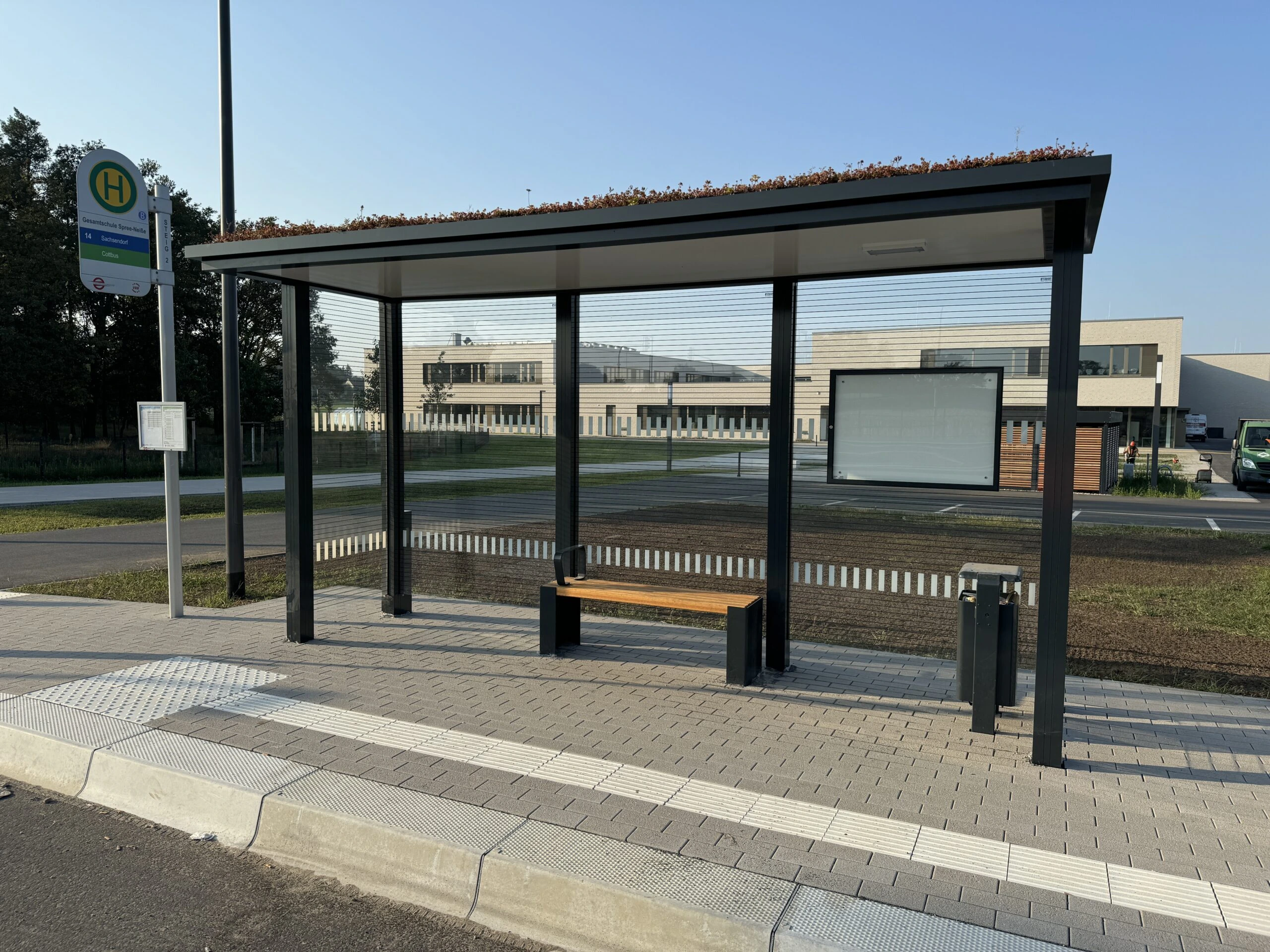 Wartehalle mit Gründach und Sitzbank an einer Bushaltestation vor einem größeren Gebäude.