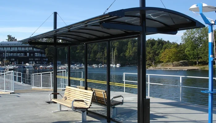 Wartehalle mit Überdachung und zwei Sitzbänken am Wasser, umgeben von Pflanzen.