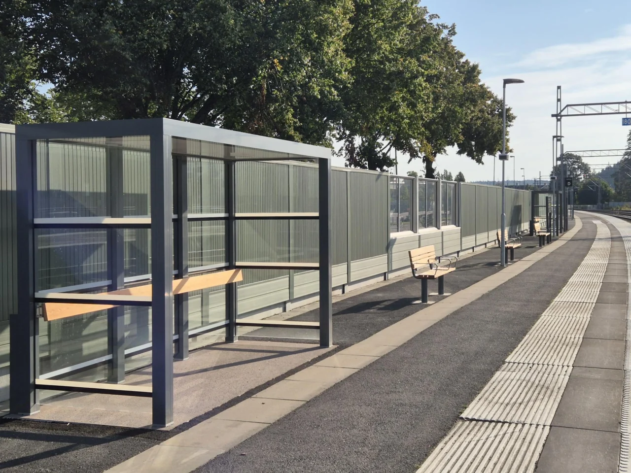 Wartehalle mit Überdachung und mehreren Sitzbänken an einem Bahnsteig, Bäume im Hintergrund.